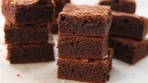 A stack of rich, fudgy chocolate brownies on a light-colored surface. The brownies are freshly cut into square pieces, showcasing their moist and dense texture. More brownie squares are visible in the background.