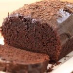 A chocolate loaf cake drizzled with chocolate glaze sits on a white rectangular plate. One slice has been cut from the front, revealing the moist and rich texture inside. The cake is garnished with chocolate shavings on top.