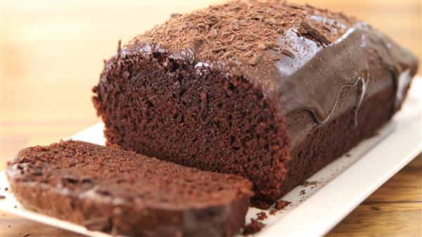 A chocolate loaf cake drizzled with chocolate glaze sits on a white rectangular plate. One slice has been cut from the front, revealing the moist and rich texture inside. The cake is garnished with chocolate shavings on top.