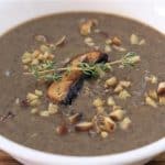 A white bowl filled with creamy mushroom soup garnished with chopped nuts, a mushroom slice, and a sprig of thyme, placed on a wooden surface.