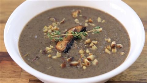 A white bowl filled with creamy mushroom soup garnished with chopped nuts, a mushroom slice, and a sprig of thyme, placed on a wooden surface.