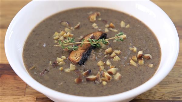 A white bowl filled with creamy mushroom soup garnished with chopped nuts, a mushroom slice, and a sprig of thyme, placed on a wooden surface.