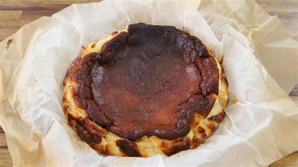 A burnt Basque cheesecake sits on a piece of parchment paper. The cheesecake features a characteristically dark, nearly burnt top layer and a slightly golden, uneven crust. The parchment paper is lightly crinkled around the cheesecake's edges.