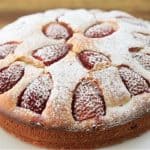 A round strawberry cake is displayed on a white plate. The cake has a light dusting of powdered sugar on top, with strawberry halves baked into the surface. The background is a wooden table.