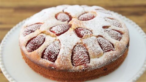 A round strawberry cake is displayed on a white plate. The cake has a light dusting of powdered sugar on top, with strawberry halves baked into the surface. The background is a wooden table.