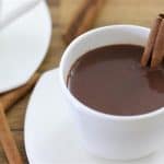 A white cup filled with hot chocolate garnished with a cinnamon stick, sitting on a matching white saucer. A few cinnamon sticks are scattered on the wooden table in the background, and a second white cup and saucer are partially visible.