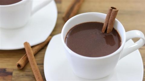 A white cup filled with hot chocolate garnished with a cinnamon stick, sitting on a matching white saucer. A few cinnamon sticks are scattered on the wooden table in the background, and a second white cup and saucer are partially visible.