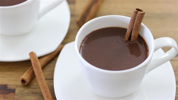 A white cup filled with hot chocolate garnished with a cinnamon stick, sitting on a matching white saucer. A few cinnamon sticks are scattered on the wooden table in the background, and a second white cup and saucer are partially visible.