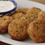 A white plate holds several falafel patties, golden brown and crispy, arranged in a semi-circle. Behind the falafel, there is a small bowl with a blue and white pattern filled with creamy white dipping sauce. The wooden table background is partially visible.