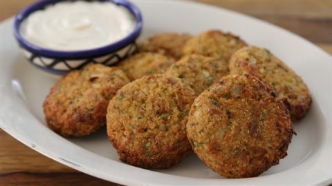 A white plate holds several falafel patties, golden brown and crispy, arranged in a semi-circle. Behind the falafel, there is a small bowl with a blue and white pattern filled with creamy white dipping sauce. The wooden table background is partially visible.