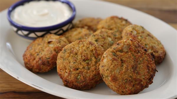 A white plate holds several falafel patties, golden brown and crispy, arranged in a semi-circle. Behind the falafel, there is a small bowl with a blue and white pattern filled with creamy white dipping sauce. The wooden table background is partially visible.