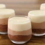 Five clear glasses filled with layered chocolate mousse are placed on a wooden surface. The dessert consists of three layers: a dark chocolate layer at the bottom, a milk chocolate layer in the middle, and a white chocolate layer on top.