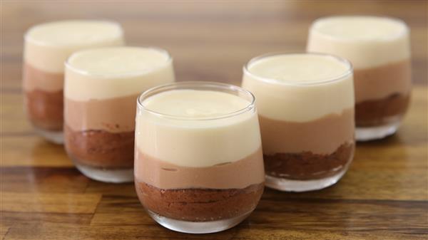 Five clear glasses filled with layered chocolate mousse are placed on a wooden surface. The dessert consists of three layers: a dark chocolate layer at the bottom, a milk chocolate layer in the middle, and a white chocolate layer on top.