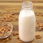 A clear glass bottle filled with almond milk stands on a wooden surface. A small glass bowl filled with raw almonds is next to the bottle, with several almonds scattered around both the bowl and the bottle on the table.