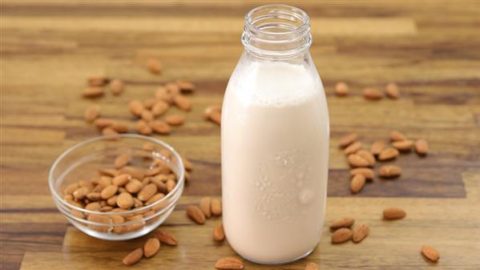 A clear glass bottle filled with almond milk stands on a wooden surface. A small glass bowl filled with raw almonds is next to the bottle, with several almonds scattered around both the bowl and the bottle on the table.