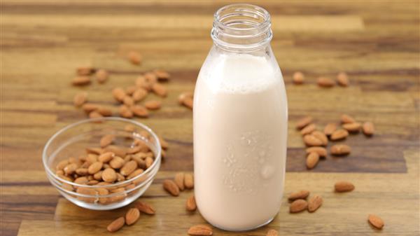 A clear glass bottle filled with almond milk stands on a wooden surface. A small glass bowl filled with raw almonds is next to the bottle, with several almonds scattered around both the bowl and the bottle on the table.