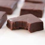 Close-up of several dark chocolate fudge pieces on white parchment paper. One piece in the foreground has a bite taken out of it, showing a smooth and creamy interior. The fudge is glossy and rich in color, arranged in a grid pattern.