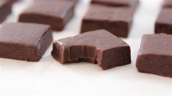 Close-up of several dark chocolate fudge pieces on white parchment paper. One piece in the foreground has a bite taken out of it, showing a smooth and creamy interior. The fudge is glossy and rich in color, arranged in a grid pattern.