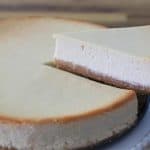 A creamy cheesecake with a golden-brown crust is shown. A slice of cheesecake is being lifted out of the whole cheesecake using a pie server, revealing the rich, smooth texture of the dessert. The background is a wooden table.