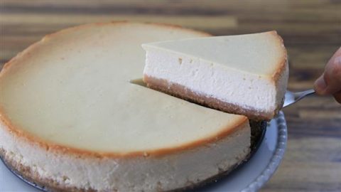 A creamy cheesecake with a golden-brown crust is shown. A slice of cheesecake is being lifted out of the whole cheesecake using a pie server, revealing the rich, smooth texture of the dessert. The background is a wooden table.