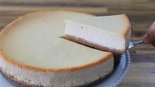 A creamy cheesecake with a golden-brown crust is shown. A slice of cheesecake is being lifted out of the whole cheesecake using a pie server, revealing the rich, smooth texture of the dessert. The background is a wooden table.