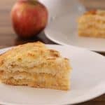 A slice of layered apple cake sits on a white plate, showcasing its moist, textured layers. In the background, another slice and a couple of whole apples are visible on a wooden table, indicating a fresh and inviting dessert setting.