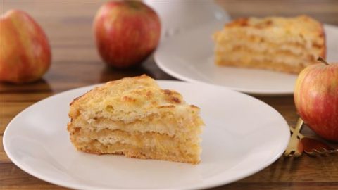 A slice of layered apple cake sits on a white plate, showcasing its moist, textured layers. In the background, another slice and a couple of whole apples are visible on a wooden table, indicating a fresh and inviting dessert setting.