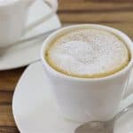 Two white ceramic mugs, each on a matching saucer with a silver teaspoon placed beside them, contain cappuccinos dusted with powdered sugar. The cups are set on a wooden table, and the focus is on the mug in the foreground.