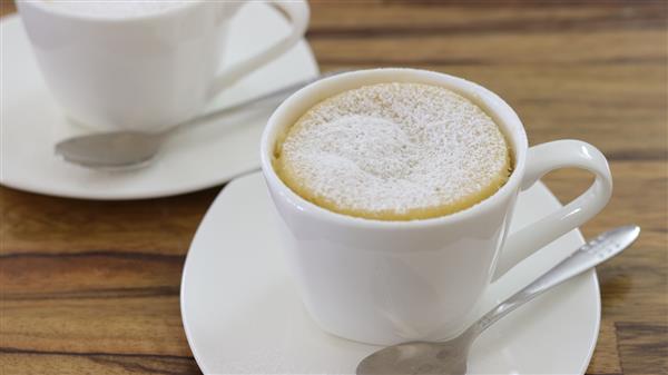 Two white ceramic mugs, each on a matching saucer with a silver teaspoon placed beside them, contain cappuccinos dusted with powdered sugar. The cups are set on a wooden table, and the focus is on the mug in the foreground.