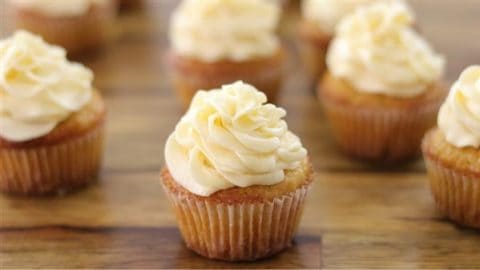 A close-up of several vanilla cupcakes with swirled cream-colored frosting arranged on a wooden surface. Each cupcake appears moist and freshly baked, topped with generous, fluffy swirls of icing. The background shows more similarly decorated cupcakes slightly out of focus.