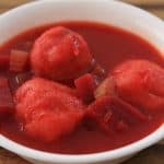 A white bowl filled with a vibrant red soup containing chunks of vegetables and what appear to be meatballs, placed on a wooden surface.