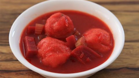 A white bowl filled with a vibrant red soup containing chunks of vegetables and what appear to be meatballs, placed on a wooden surface.