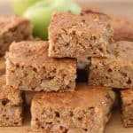 A stack of homemade apple cinnamon bars sits on a wooden surface. The top bar reveals moist texture with visible apple chunks. In the background, a blurred green apple adds to the rustic and fresh feel of the image.