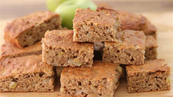 A stack of homemade apple cinnamon bars sits on a wooden surface. The top bar reveals moist texture with visible apple chunks. In the background, a blurred green apple adds to the rustic and fresh feel of the image.