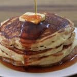 A stack of five golden-brown pancakes on a white plate, topped with a melting pat of butter. Syrup is being drizzled over the top, cascading down the sides of the pancakes. The background is a wooden table surface.