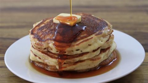 A stack of five golden-brown pancakes on a white plate, topped with a melting pat of butter. Syrup is being drizzled over the top, cascading down the sides of the pancakes. The background is a wooden table surface.