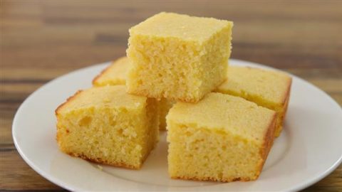 Five pieces of freshly baked, golden cornbread stacked on a white plate against a wooden background. The cornbread squares have a moist and fluffy texture with slightly browned edges.