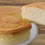 A light and fluffy Japanese cheesecake is shown on a white plate. A single slice is being lifted out with a cake server, revealing the cake's airy texture. The background is a wooden surface.