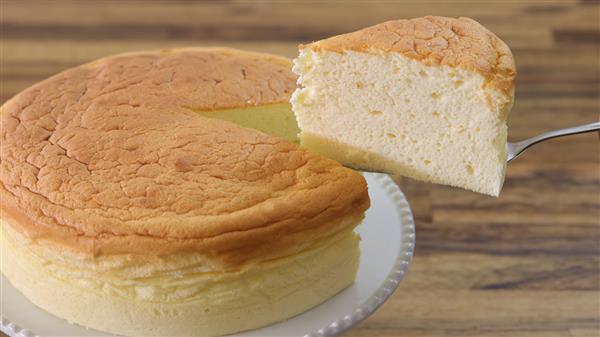 A light and fluffy Japanese cheesecake is shown on a white plate. A single slice is being lifted out with a cake server, revealing the cake's airy texture. The background is a wooden surface.