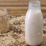 A glass bottle filled with oat milk is placed on a wooden table. Next to it is a small glass jar filled with rolled oats. Loose rolled oats are scattered on the table around both the bottle and the jar.