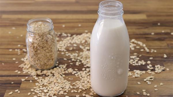 A glass bottle filled with oat milk is placed on a wooden table. Next to it is a small glass jar filled with rolled oats. Loose rolled oats are scattered on the table around both the bottle and the jar.