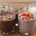 Five glasses of chia seed pudding arranged on a wooden table. Each glass features a different flavor and topping combination, including chocolate with bananas, chocolate with almonds, vanilla with berries, vanilla with walnuts and strawberries, and plain vanilla.