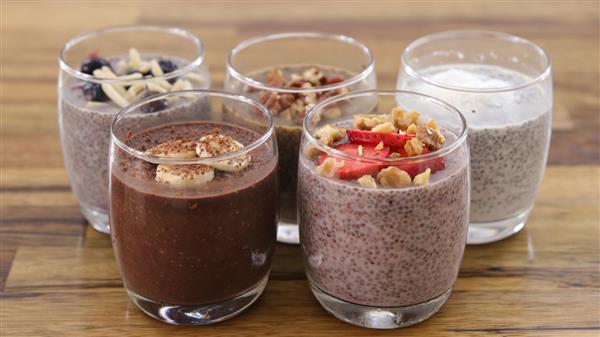 Five glasses of chia seed pudding arranged on a wooden table. Each glass features a different flavor and topping combination, including chocolate with bananas, chocolate with almonds, vanilla with berries, vanilla with walnuts and strawberries, and plain vanilla.