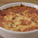 A round, white casserole dish containing a baked dish with a golden-brown, bubbly, melted cheese topping. The dish is placed on a wooden surface with a round wooden trivet beneath it.