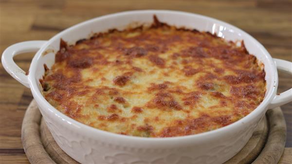 A round, white casserole dish containing a baked dish with a golden-brown, bubbly, melted cheese topping. The dish is placed on a wooden surface with a round wooden trivet beneath it.