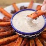 A hand dips a roasted carrot stick into a small bowl of creamy dipping sauce. The bowl is centered on a plate arranged with more roasted carrot sticks, all of which are crispy and seasoned. The background is a blurred wooden surface.