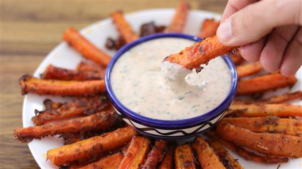 A hand dips a roasted carrot stick into a small bowl of creamy dipping sauce. The bowl is centered on a plate arranged with more roasted carrot sticks, all of which are crispy and seasoned. The background is a blurred wooden surface.