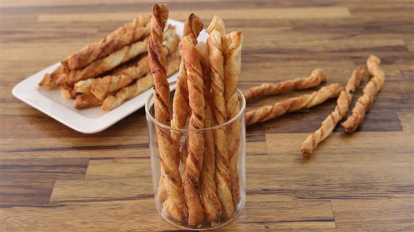 A transparent glass cup holds multiple twisted breadsticks on a wooden surface. In the background, a rectangular white plate is filled with more breadsticks, and several breadsticks are scattered on the table.