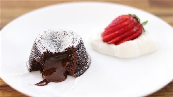 A delicious chocolate lava cake with molten center is served on a white plate. The cake is dusted with powdered sugar and is accompanied by a dollop of whipped cream topped with a sliced strawberry.