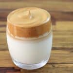 A clear glass of dalgona coffee with a layer of whipped coffee foam on top of milk. The background surface is a wooden table.